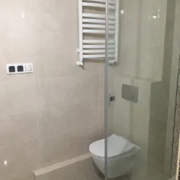 Nowoczesna łazienka z beżowymi płytkami, wiszącą toaletą, grzejnikiem drabinkowym i szklaną kabiną prysznicową.