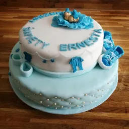 Dwupoziomowy tort w odcieniach błękitu z napisem 'Święty Ernesta', dekorowany figurkami niemowląt w gnieździe z kwiatów, małymi bucikami i ubrankami.