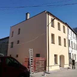 Odnowiona elewacja budynku w odcieniach beżu, widoczna drabina oparta o ścianę, czerwony samochód dostawczy w cieniu oraz pracownik zamiatający chodnik przed budynkiem.