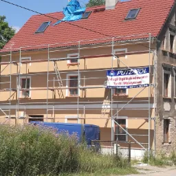 Remont elewacji w Nowej Rudzie: dom w rusztowaniach, część budynku otynkowana na beżowo, druga część z odsłoniętą cegłą, na dachu niebieska plandeka, reklama tynków maszynowych na rusztowaniu.