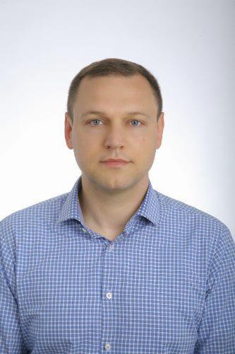 Portret mężczyzny w błękitnej koszuli w drobną kratkę na jasnym tle.