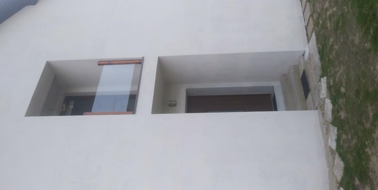 Elewacja budynku z minimalistycznym oknem z drewnianą ramą i zagłębionym wejściem z drzwiami, widoczne schody i fragment trawnika.