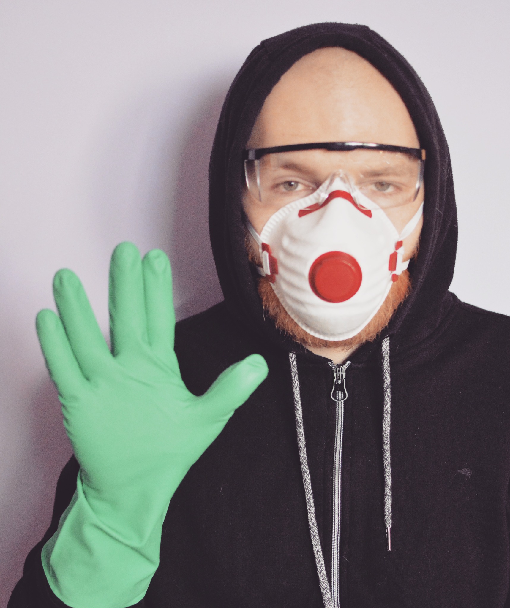 Mężczyzna w kapturze, masce przeciwpyłowej i okularach ochronnych, w zielonej rękawicy ochronnej, wyciąga dłoń z rozwartymi palcami.