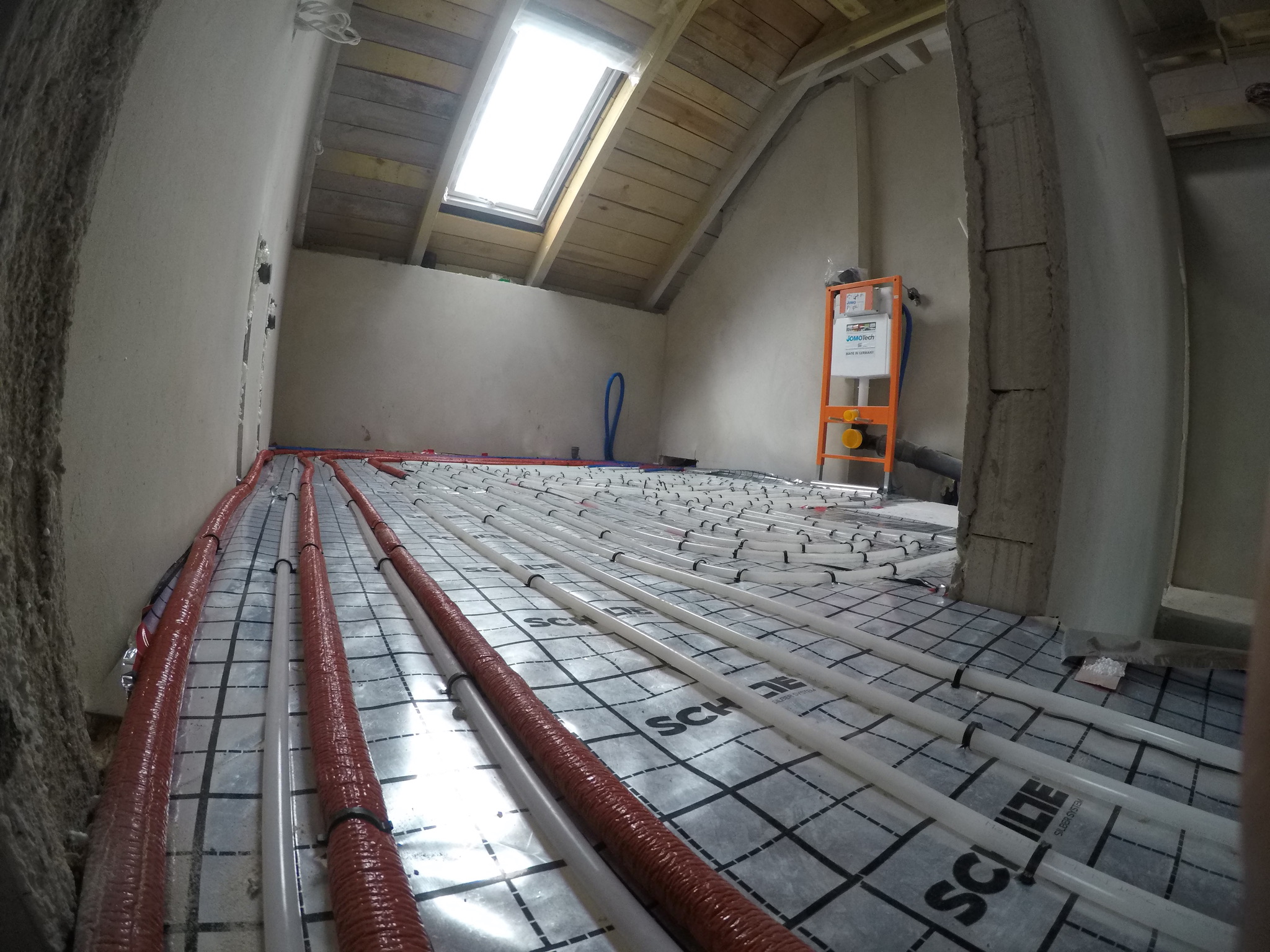 Instalacja ogrzewania podłogowego z rurami w izolacji, widok z perspektywy podłogi, w tle okno dachowe i elementy instalacji sanitarnej.