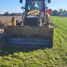 Żółta koparko-ładowarka CAT z brudną łyżką stoi na trawiastym terenie, w tle świeżo przekopana ziemia i błękitne niebo.