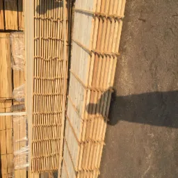 Stosy drewnianych desek i elementów konstrukcyjnych, częściowo owiniętych folią, ułożone na placu budowy w pobliżu ściany budynku z oknami.