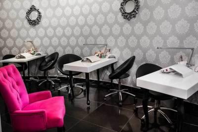 Wnętrze gabinetu kosmetycznego z trzema stanowiskami do manicure, każdy z lampą i krzesłem, na ścianie ozdobne lustra i tapeta we wzory, na pierwszym planie różowy fotel.