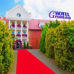 Elegancki budynek hotelu w Sękocinie Starym z czerwonym dywanem prowadzącym do wejścia, otoczony zielenią i podświetlonym szyldem 'Hotel Groman'.