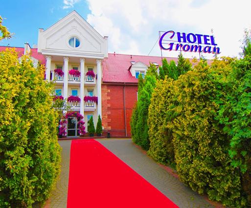 Elegancki budynek hotelu w Sękocinie Starym z czerwonym dywanem prowadzącym do wejścia, otoczony zielenią i podświetlonym szyldem 'Hotel Groman'.