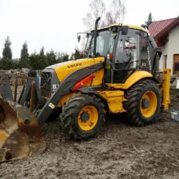 Żółto-szara koparko-ładowarka Volvo BL71 na błotnistym terenie budowy, z domem w tle i elementami konstrukcyjnymi w otoczeniu.
