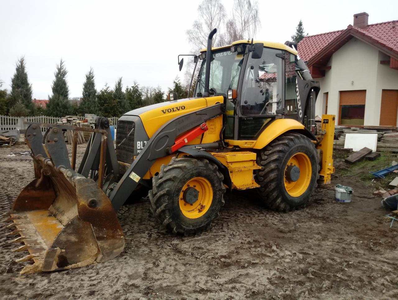 Żółto-szara koparko-ładowarka Volvo BL71 na błotnistym terenie budowy, z domem w tle i elementami konstrukcyjnymi w otoczeniu.