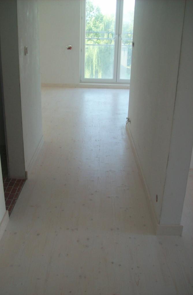 Jasna, drewniana podłoga w nowym mieszkaniu, widok na okno balkonowe i fragment ściany z listwą przypodłogową.