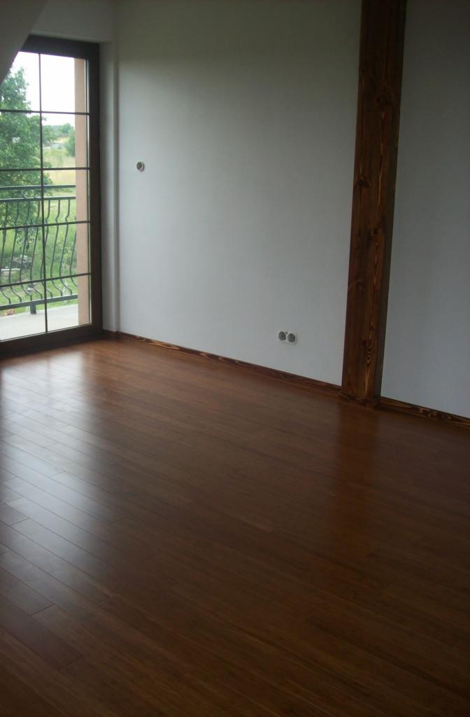 Ciemnobrązowa podłoga z paneli, widok na balkon z metalową balustradą i zielenią na zewnątrz, fragment drewnianej kolumny przy ścianie.