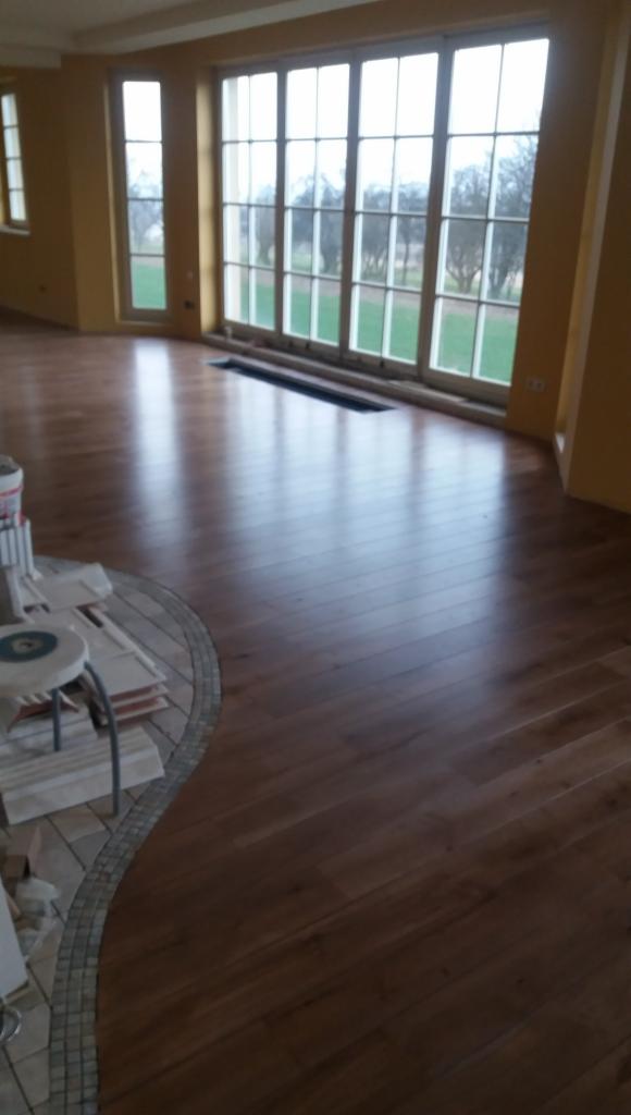 Błyszcząca, nowo ułożona podłoga z paneli drewnianych w przestronnym salonie z dużymi oknami, widoczne materiały wykończeniowe i fragment mozaiki podłogowej.