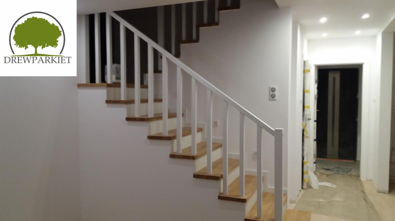 Nowoczesne schody drewniane z białymi balustradami i drewnianymi stopniami, widoczne logo firmy na ścianie, w tle korytarz z zabezpieczonymi drzwiami.