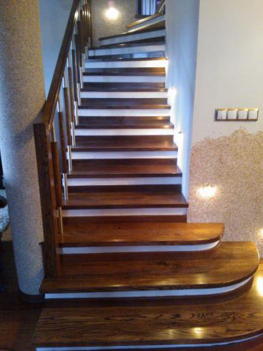 Kręte schody z ciemnego drewna z białymi akcentami, widoczne z dołu, z kolumną obok i oświetleniem w tle.