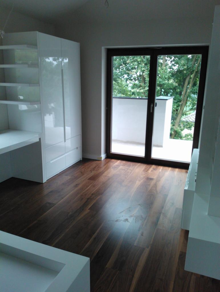 Ciemne panele podłogowe ułożone w pokoju z białymi meblami i balkonem z widokiem na zieleń.