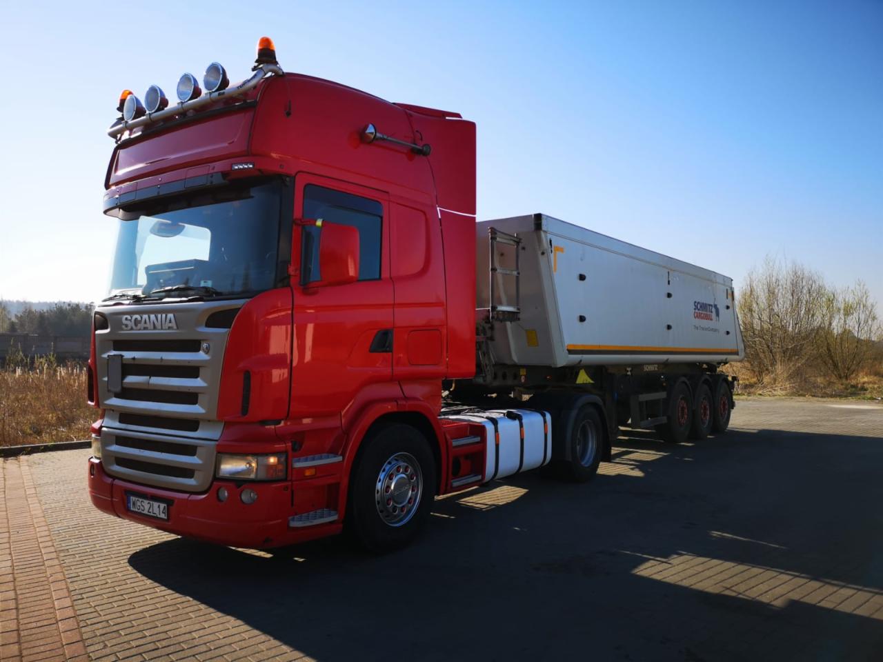 Czerwony ciągnik siodłowy Scania z naczepą typu wywrotka marki Schmitz Cargobull na parkingu, widoczne światła ostrzegawcze na dachu kabiny.
