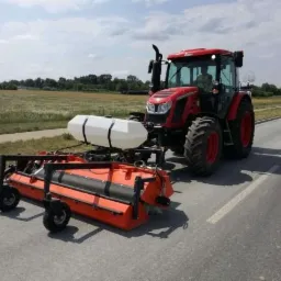 Czerwony traktor z pomarańczową szczotką czyszczącą pobocze drogi asfaltowej w słoneczny dzień.