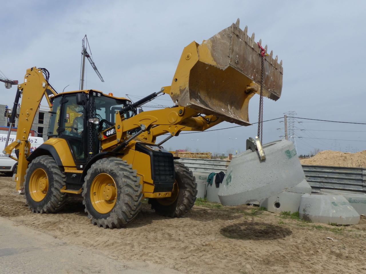Żółta koparko-ładowarka podnosi betonowy element studni za pomocą łańcucha na placu budowy, w tle widać dźwigi i budynki.