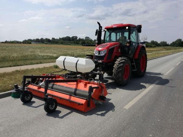 Czerwony traktor z pomarańczową szczotką czyszczącą pobocze drogi asfaltowej w słoneczny dzień.