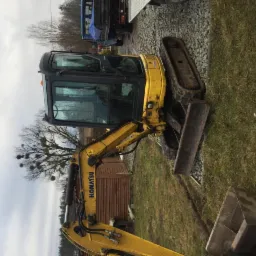 Żółta minikoparka Komatsu na trawiastym terenie z widocznym łyżką, gąsienicami i kabiną, zaparkowana obok żwirowej drogi i drewnianego budynku w tle.