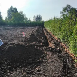 Wykopany rów w Grajewie, widoczne ślady opon i fragment białej rury, w tle zieleń drzew.