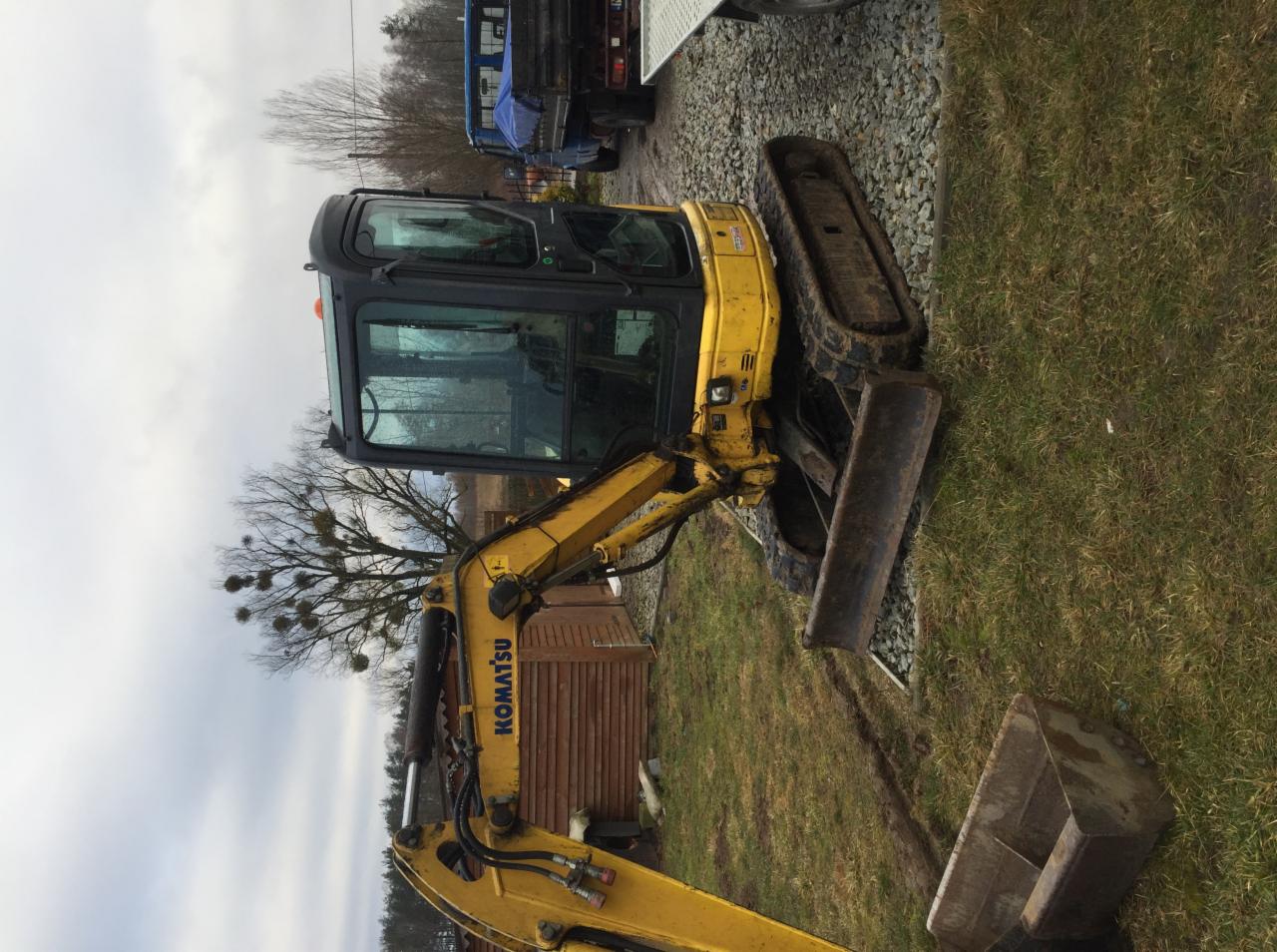 Żółta minikoparka Komatsu na trawiastym terenie z widocznym łyżką, gąsienicami i kabiną, zaparkowana obok żwirowej drogi i drewnianego budynku w tle.