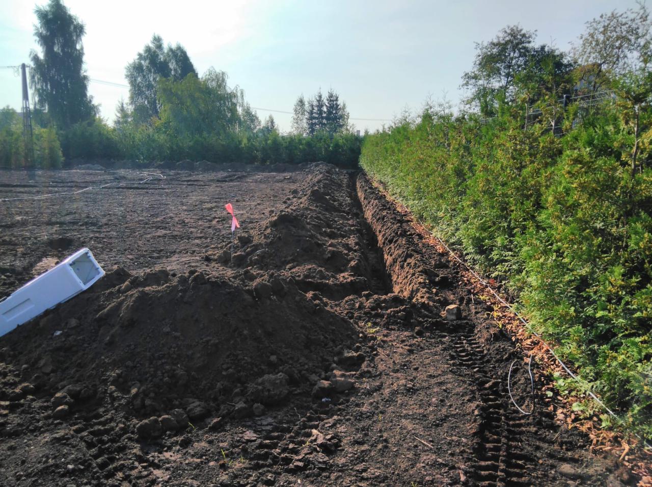 Wykopany rów w Grajewie, widoczne ślady opon i fragment białej rury, w tle zieleń drzew.