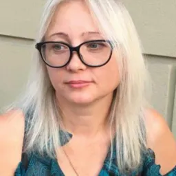 Portret kobiety w okularach, w bluzce w kratę, z platynowymi włosami, pozującej na tle ściany. Widoczny fragment ramienia i biżuterii.