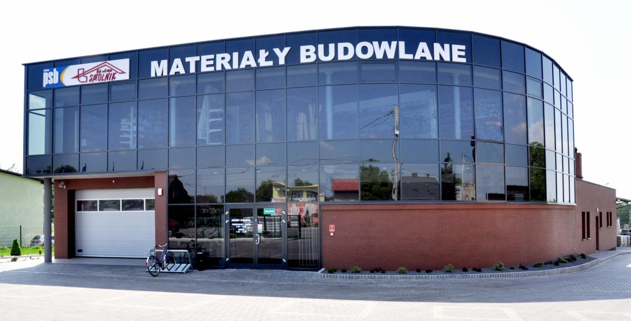 Dwukondygnacyjny budynek składu budowlanego z dużymi przeszklonymi oknami i napisem 'Materiały Budowlane', rower zaparkowany przed garażem.