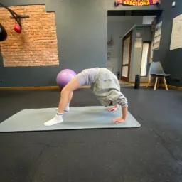 Osoba w szarej bluzie wykonuje pozycję mostka na szarej macie w sali z ceglaną ścianą i piłką do ćwiczeń, w tle widoczny szalik FC Barcelona.