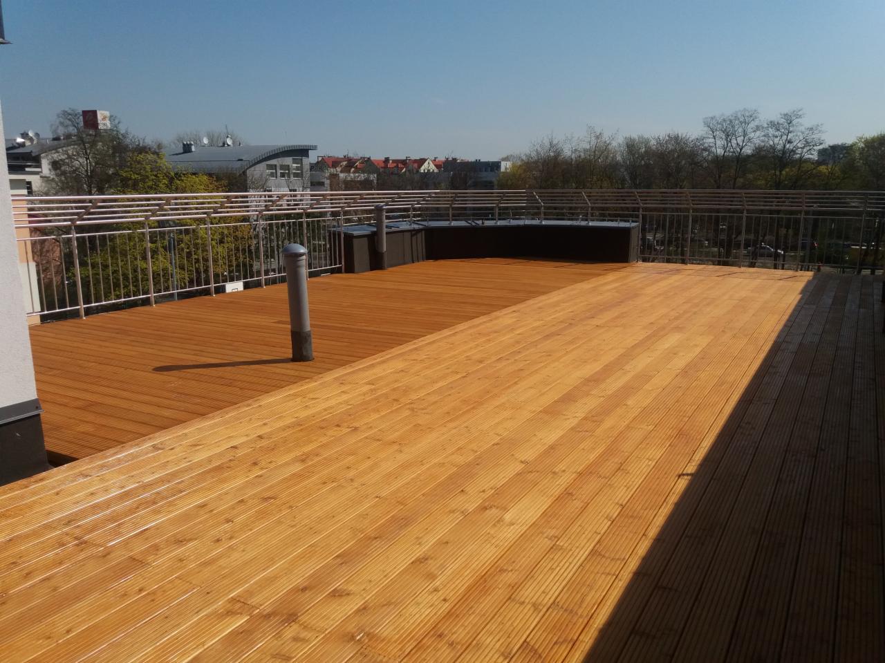Rozległy taras na dachu budynku, z drewnianą podłogą w dwóch odcieniach brązu, otoczony metalową balustradą, z widokiem na okoliczną zabudowę i drzewa w słoneczny dzień.