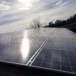Panel słoneczny na dachu budynku w pochmurny dzień, widoczne odbicie słońca na panelach i drzewa w tle.