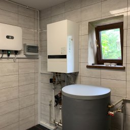 HORIZONTAL ENERGY RAFAŁ WITCZAK - Wnętrze kotłowni z białym, wiszącym kotłem, szarym zbiornikiem akumulacyjnym, falownikiem z logiem producenta oraz skrzynką elektryczną, widoczne rury i okno.