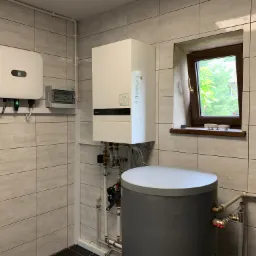 Wnętrze kotłowni z białym, wiszącym kotłem, szarym zbiornikiem akumulacyjnym, falownikiem z logiem producenta oraz skrzynką elektryczną, widoczne rury i okno.