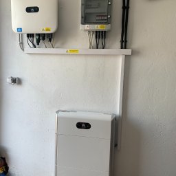 HORIZONTAL ENERGY RAFAŁ WITCZAK - Biała bateria energii i inwerter fotowoltaiczny Huawei zamontowane na pomalowanej na biało ścianie z wyeksponowanymi przewodami w peszlach i skrzynką bezpiecznikową. Podłoga z kostki brukowej.