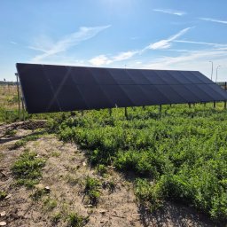 HORIZONTAL ENERGY RAFAŁ WITCZAK - Rząd paneli fotowoltaicznych na metalowych wspornikach, ustawiony na zarośniętym terenie z widokiem na błękitne niebo z chmurami.