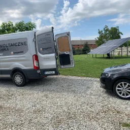 Montaż paneli fotowoltaicznych na trawniku z widocznym samochodem osobowym i busem firmy instalacyjnej z otwartymi drzwiami, w tle budynek gospodarczy.