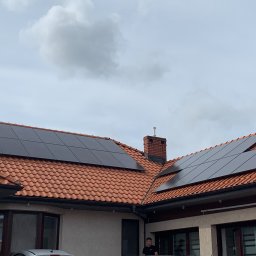 HORIZONTAL ENERGY RAFAŁ WITCZAK - Czerwony dach z dachówki ceramicznej z zamontowanymi czarnymi panelami fotowoltaicznymi, widoczny komin z cegły i fragment elewacji domu z oknami.