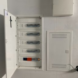 HORIZONTAL ENERGY RAFAŁ WITCZAK - Otwarta biała skrzynka rozdzielcza z bezpiecznikami na kafelkowanej ścianie, obok zamknięta skrzynka z wentylacją i dodatkowa biała obudowa na górze.