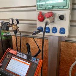 HORIZONTAL ENERGY RAFAŁ WITCZAK - Miernik Sonel MPI-540-PV podłączony do tablicy rozdzielczej Elektromet z gniazdami siłowymi, obok butla z gazem technicznym, na tle ściany z boazerii.