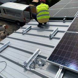 HORIZONTAL ENERGY RAFAŁ WITCZAK - Montaż paneli słonecznych na metalowym dachu: instalator w kamizelce, części montażowe, samochód dostawczy z napisem 'Fotowoltaiczne' i betoniarka w tle.