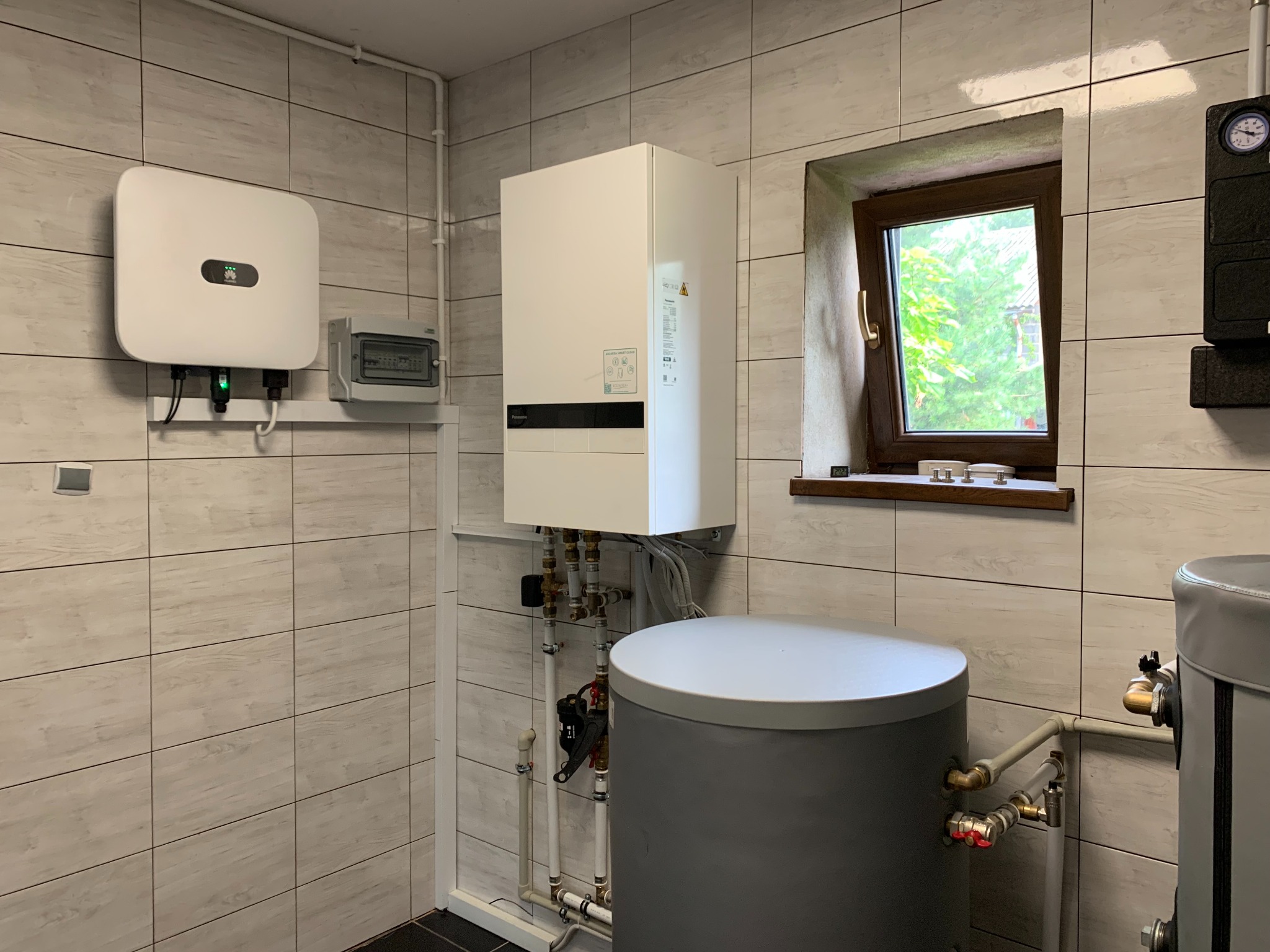 Wnętrze kotłowni z białym, wiszącym kotłem, szarym zbiornikiem akumulacyjnym, falownikiem z logiem producenta oraz skrzynką elektryczną, widoczne rury i okno.