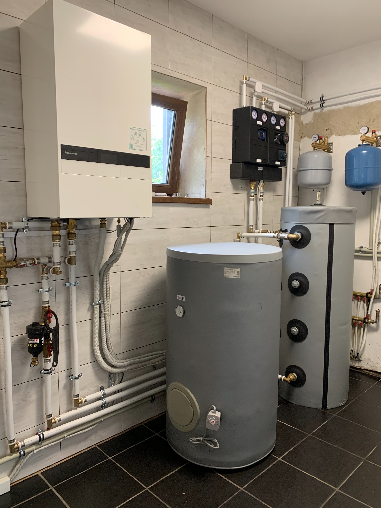 Biała jednostka wewnętrzna pompy ciepła Panasonic Aquarea Smart Cloud, dwa szare zbiorniki akumulacyjne ciepłej wody z izolacją, czarna skrzynka sterownicza z manometrami, instalacja hydrauliczna...