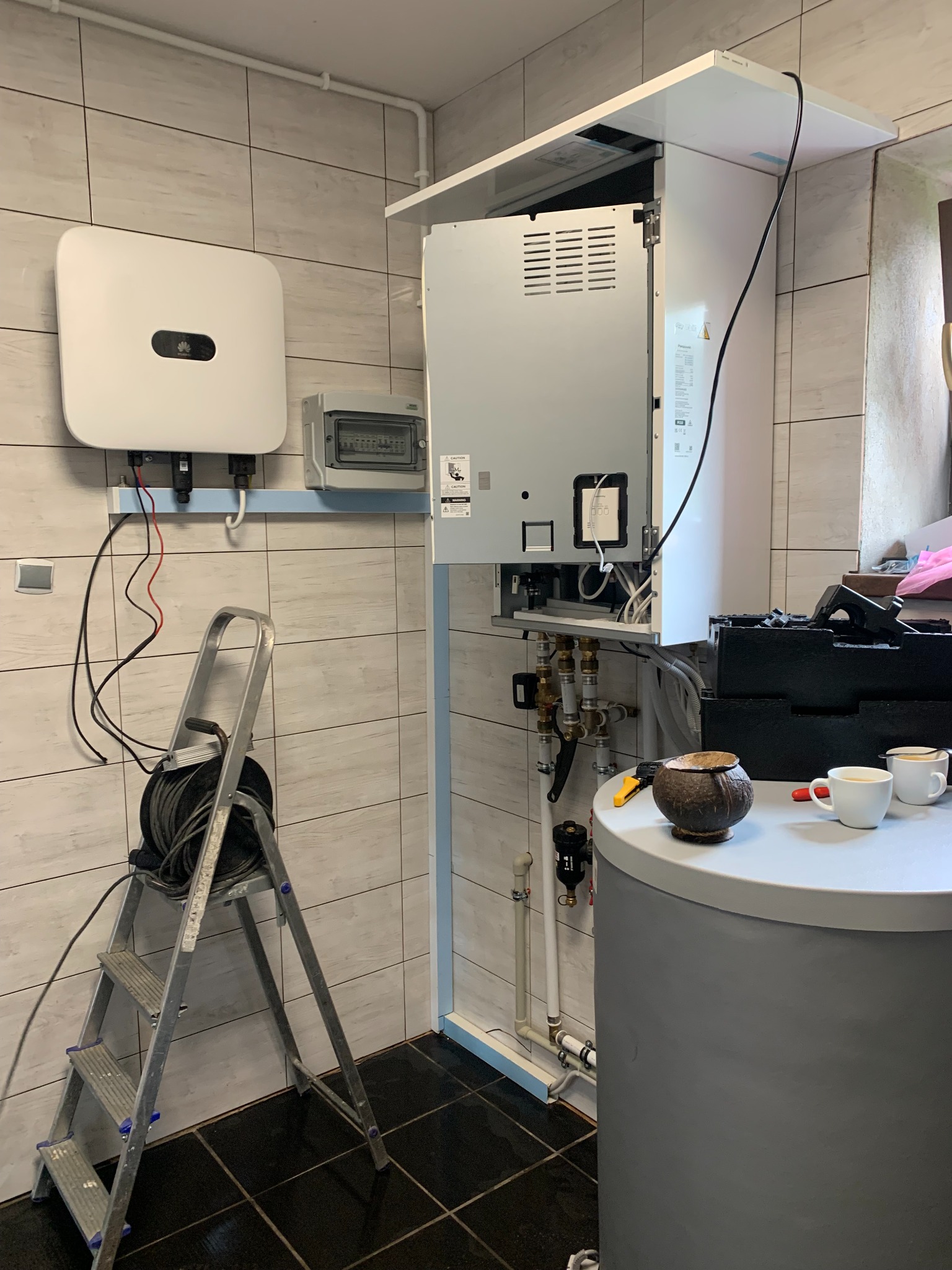 Wnętrze kotłowni z zamontowanym inwerterem fotowoltaicznym Huawei, rozdzielnicą elektryczną, drabinką z kablem, zbiornikiem akumulacyjnym i dwoma kubkami na blacie.