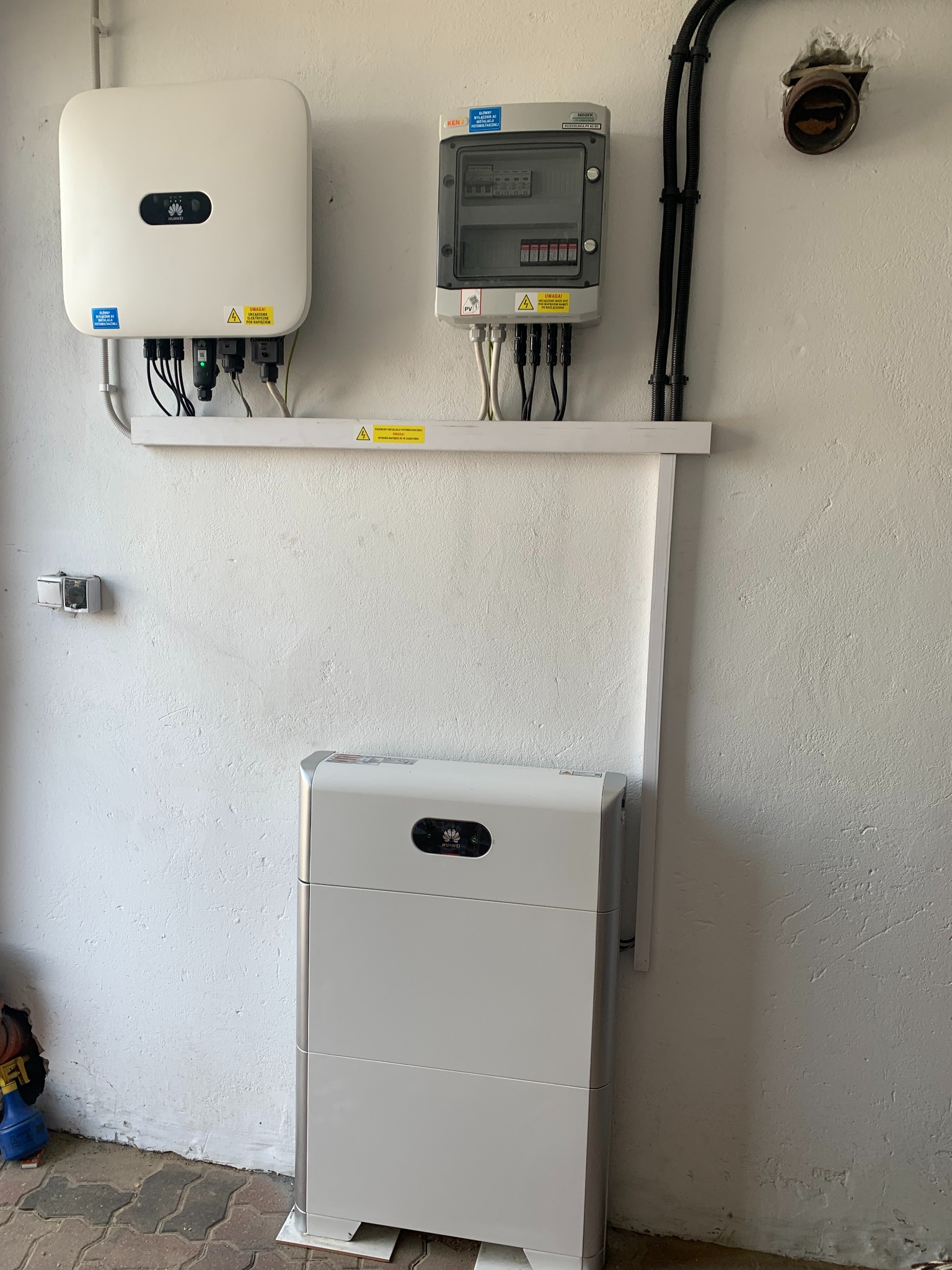 Biała bateria energii i inwerter fotowoltaiczny Huawei zamontowane na pomalowanej na biało ścianie z wyeksponowanymi przewodami w peszlach i skrzynką bezpiecznikową. Podłoga z kostki brukowej.