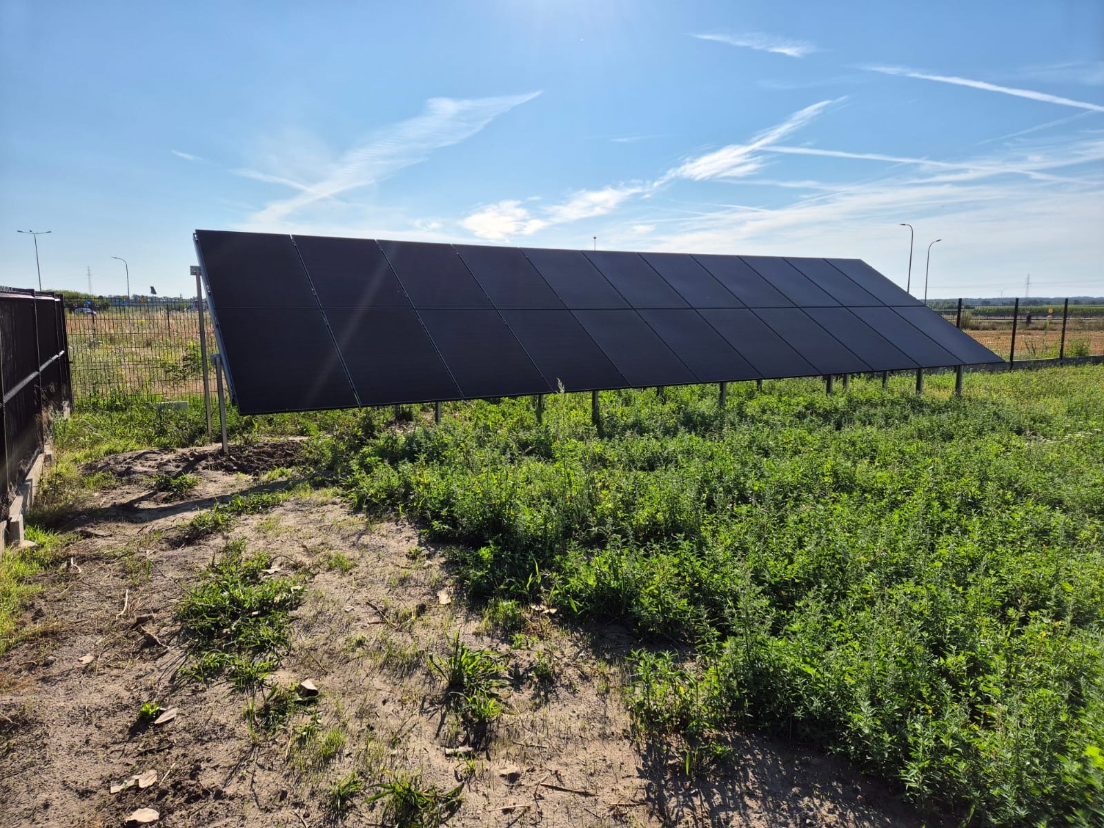Rząd paneli fotowoltaicznych na metalowych wspornikach, ustawiony na zarośniętym terenie z widokiem na błękitne niebo z chmurami.