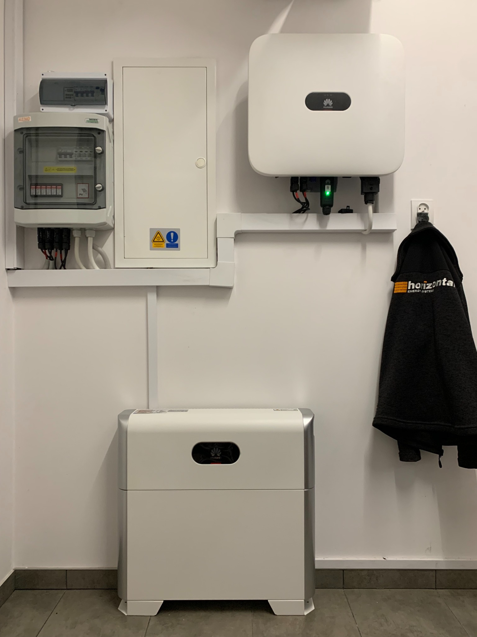 Biała ściana z zamontowanym falownikiem, skrzynką elektryczną, akumulatorem energii i wiszącą kurtką firmową.