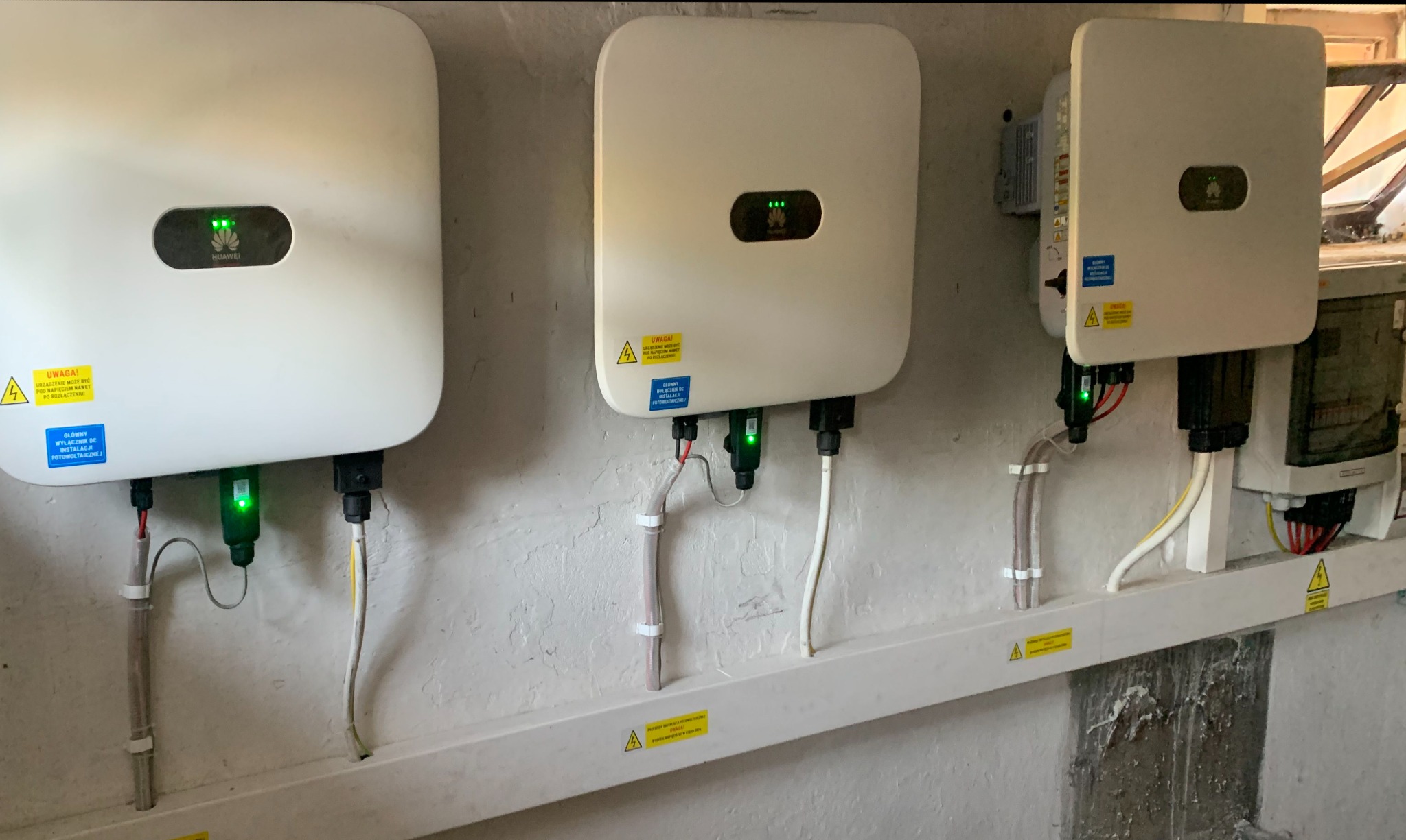 Trzy białe falowniki marki Huawei zamontowane na ścianie, połączone przewodami elektrycznymi zabezpieczonymi listwą przypodłogową. Widoczne naklejki ostrzegawcze o napięciu i wyłączniku DC...